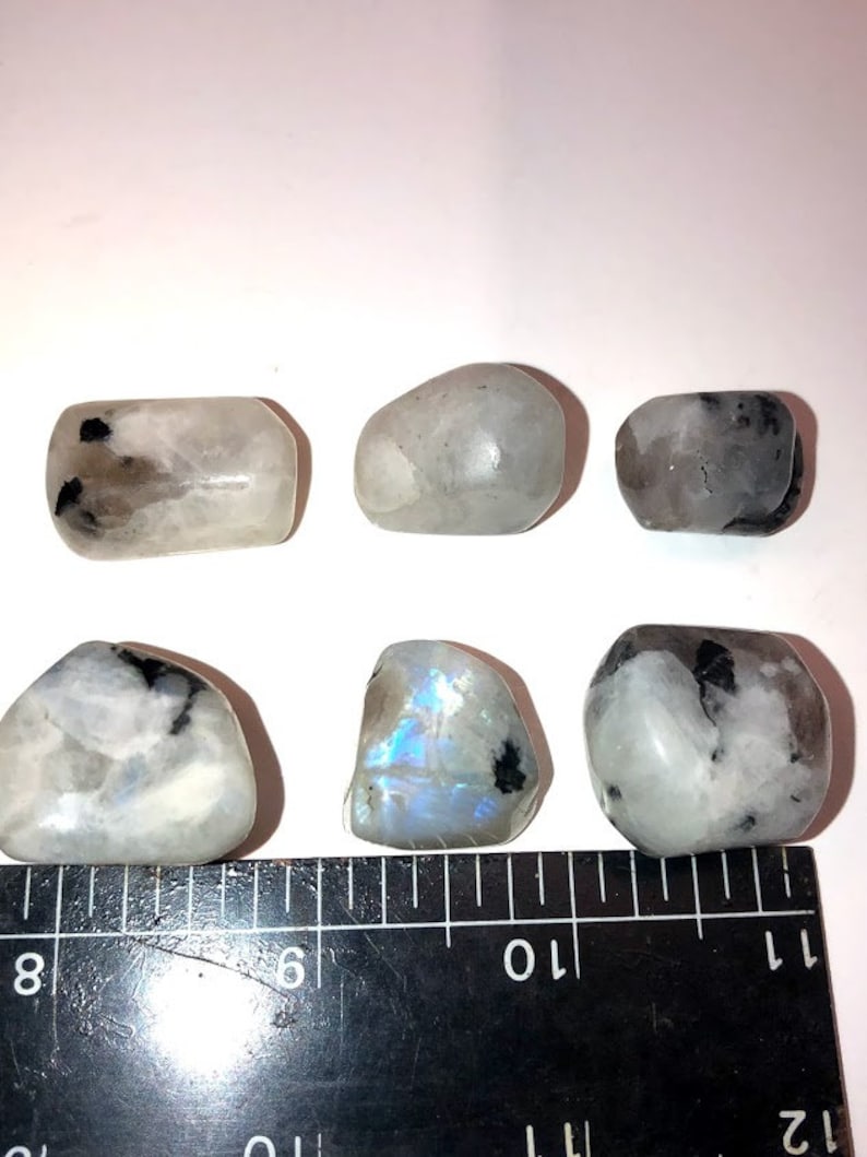 May include: Six smooth, white moonstone tumbled stones with black inclusions. The stones are arranged in two rows of three, with a ruler in the bottom of the image for size reference.