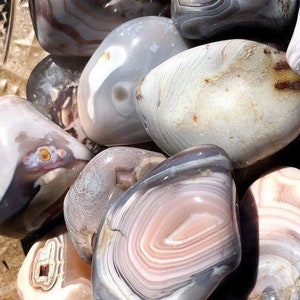 May include: A collection of polished agate stones with various patterns and colors, including white, gray, and pink. The stones are smooth and have a natural, organic look.
