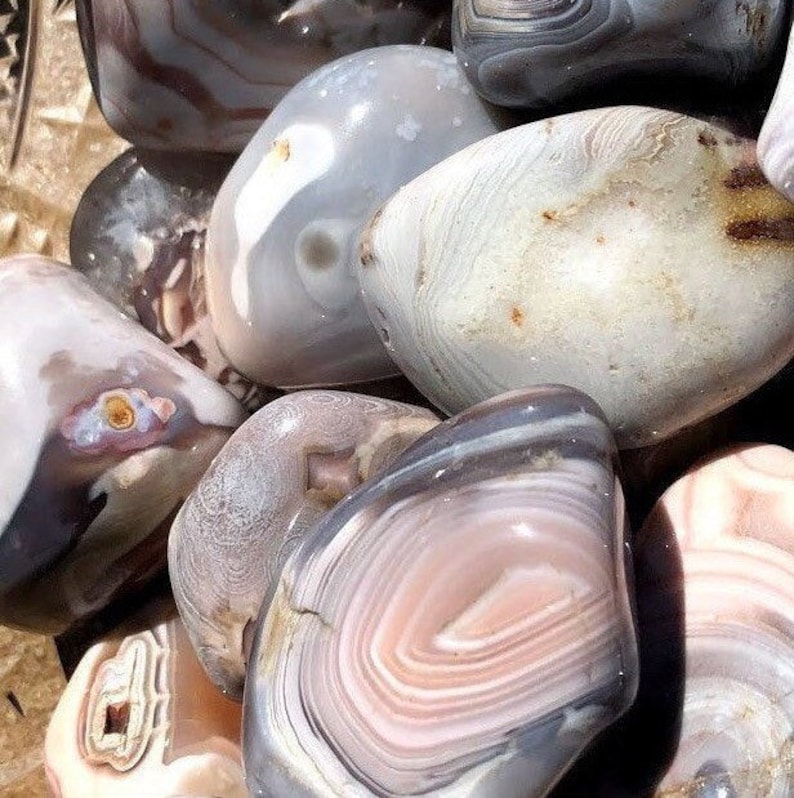May include: A collection of polished agate stones with various patterns and colors, including white, gray, and pink. The stones are smooth and have a natural, organic look.