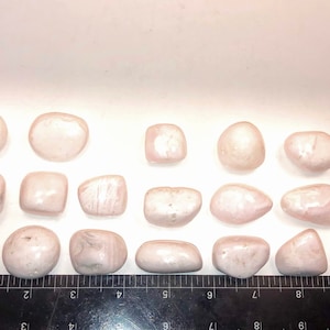 Op de afbeelding: Een verzameling van 17 gladde, gepolijste roze en witte stenen. De stenen zijn van verschillende vormen en maten en zijn in een rij op een wit oppervlak gerangschikt.