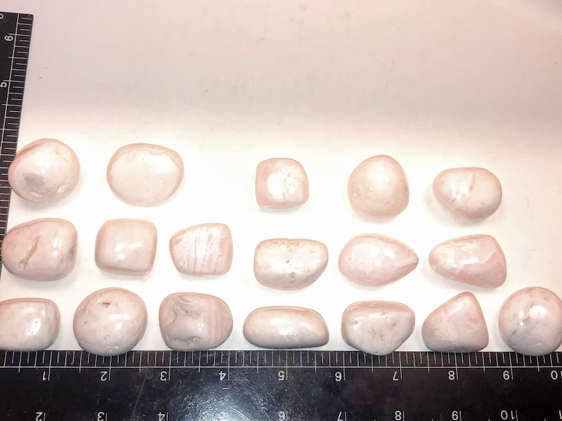 Op de afbeelding: Een verzameling van 17 gladde, gepolijste roze en witte stenen. De stenen zijn van verschillende vormen en maten en zijn in een rij op een wit oppervlak gerangschikt.