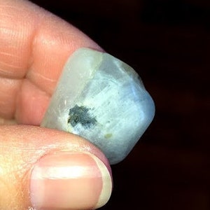 May include: A smooth, white, opaque gemstone with a dark gray inclusion. The gemstone is irregularly shaped and has a slightly rounded surface.
