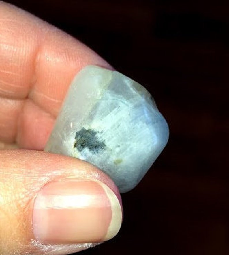 May include: A smooth, white, opaque gemstone with a dark gray inclusion. The gemstone is irregularly shaped and has a slightly rounded surface.