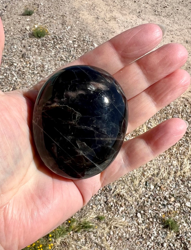 May include: A smooth, black, polished stone with a striated pattern. The stone is round and has a glossy finish.