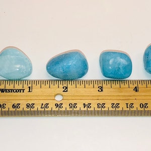 May include: Four smooth, light blue aquamarine stones of varying sizes, measured against a wooden ruler with inch markings.