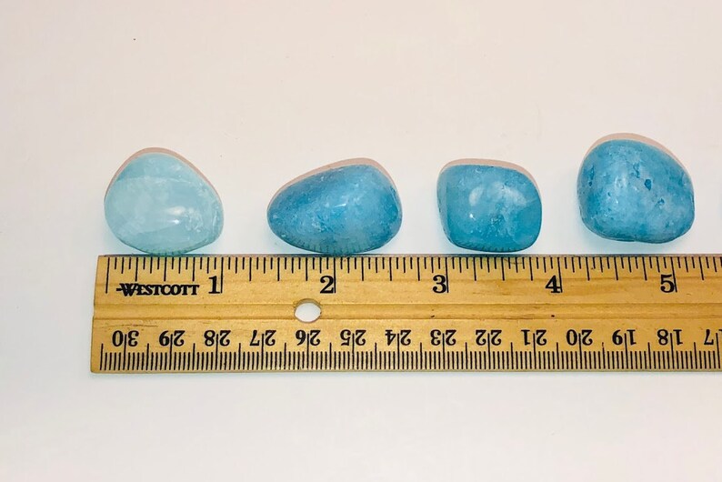 May include: Four smooth, light blue aquamarine stones of varying sizes, measured against a wooden ruler with inch markings.