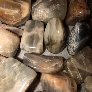 May include: A collection of smooth, polished moonstone stones in various shades of gray and white. The stones are tumbled and have a natural, organic look.