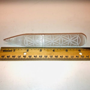 May include: A white selenite wand with a carved flower of life design. The wand is approximately 6 inches long and is measured against a wooden ruler.