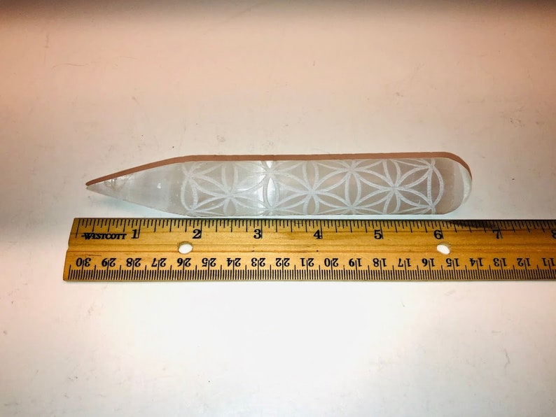 May include: A white selenite wand with a carved flower of life design. The wand is approximately 6 inches long and is measured against a wooden ruler.