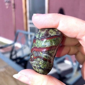 May include: A green, red, and brown polished stone with a smooth, rounded shape. The stone has a unique pattern of swirling colors and textures.