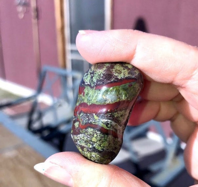 May include: A green, red, and brown polished stone with a smooth, rounded shape. The stone has a unique pattern of swirling colors and textures.