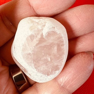 May include: A smooth, white, heart-shaped crystal with a translucent appearance. The crystal is held in a hand.