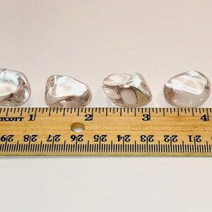 May include: Four clear quartz tumbled stones, each approximately 2.5 cm in size, are lined up on a wooden ruler. The ruler is marked with inches and fractions of an inch.