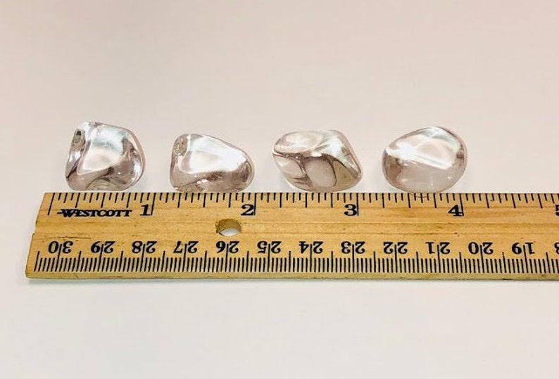 May include: Four clear quartz tumbled stones, each approximately 2.5 cm in size, are lined up on a wooden ruler. The ruler is marked with inches and fractions of an inch.