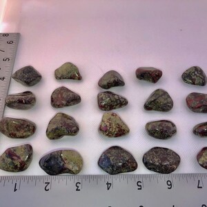 May include: Twenty tumbled stones with a mix of green, red, and brown colors. The stones are arranged in rows on a white surface with a ruler for scale.