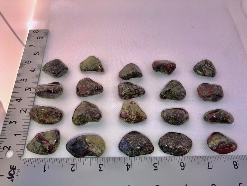 May include: Twenty tumbled stones with a mix of green, red, and brown colors. The stones are arranged in rows on a white surface with a ruler for scale.