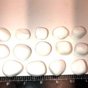 May include: A collection of 14 smooth, white, oval-shaped stones. The stones are arranged in rows on a white surface with a black ruler underneath.