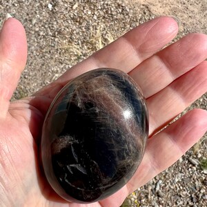 May include: A smooth, black and brown gemstone sphere with a high polish. The sphere is about the size of a golf ball.