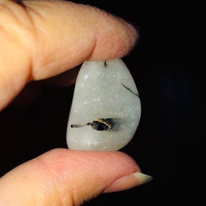 Puede incluir: Una piedra lisa, blanca y pulida con inclusiones negras y doradas. La piedra es aproximadamente del tama&ntilde;o de un huevo peque&ntilde;o.