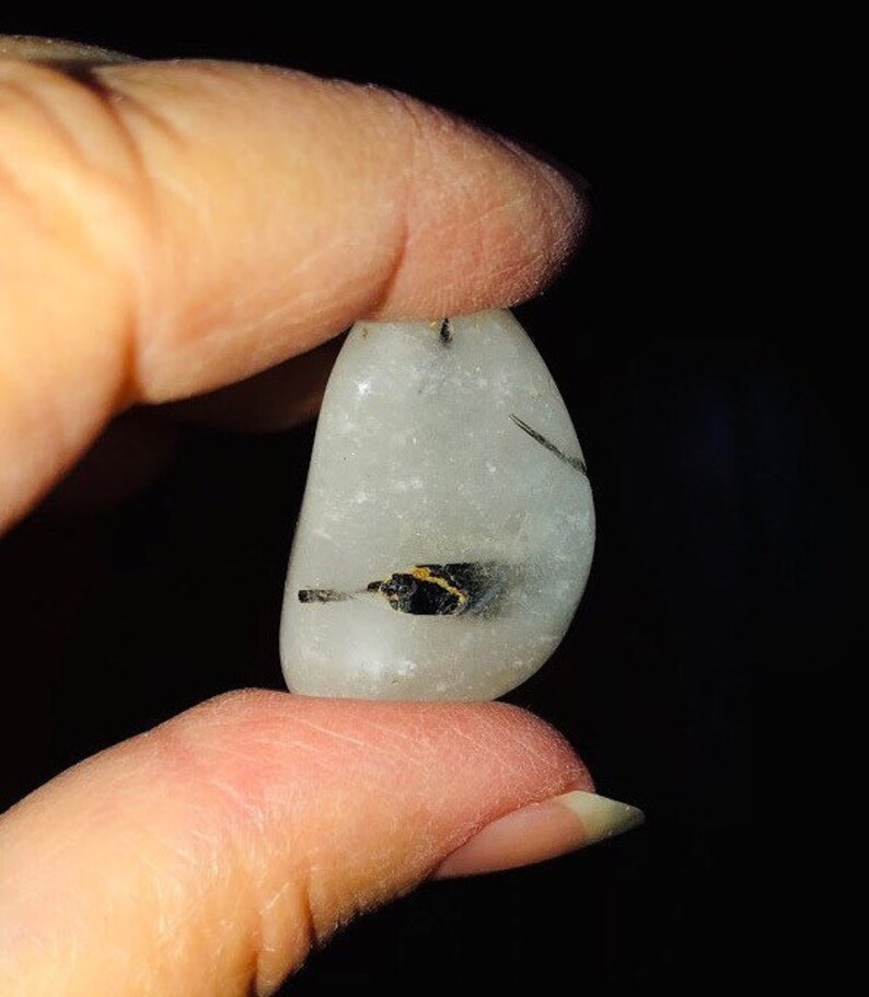 Puede incluir: Una piedra lisa, blanca y pulida con inclusiones negras y doradas. La piedra es aproximadamente del tama&ntilde;o de un huevo peque&ntilde;o.