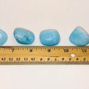May include: Four smooth, light blue aquamarine tumbled stones of varying sizes, measured against a wooden ruler.