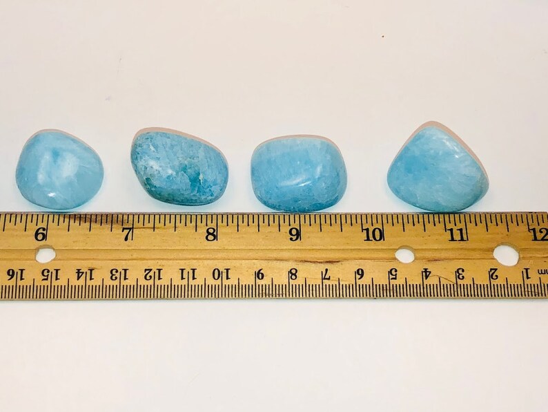 May include: Four smooth, light blue aquamarine tumbled stones of varying sizes, measured against a wooden ruler.