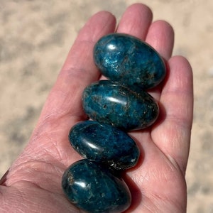 Puede incluir: Cuatro piedras de apatita azul lisas y pulidas, cada una con un tono de azul ligeramente diferente, est&aacute;n apiladas una encima de la otra en la mano de una persona.