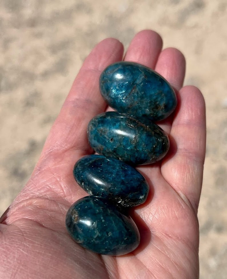 Puede incluir: Cuatro piedras de apatita azul lisas y pulidas, cada una con un tono de azul ligeramente diferente, est&aacute;n apiladas una encima de la otra en la mano de una persona.