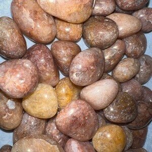 May include: A collection of tumbled rose quartz stones in various shades of pink, brown, and white. The stones are smooth and polished, with a translucent appearance.
