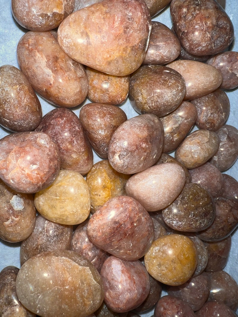 May include: A collection of tumbled rose quartz stones in various shades of pink, brown, and white. The stones are smooth and polished, with a translucent appearance.