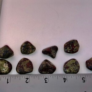 May include: Nine tumbled stones with a green, brown, and red marbled pattern. The stones are arranged in a row on a white surface with a ruler for scale.