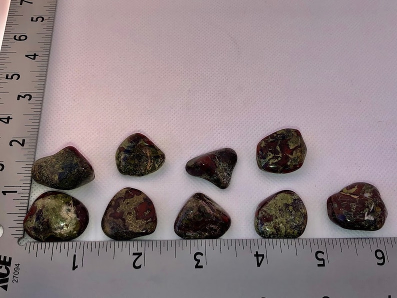 May include: Nine tumbled stones with a green, brown, and red marbled pattern. The stones are arranged in a row on a white surface with a ruler for scale.