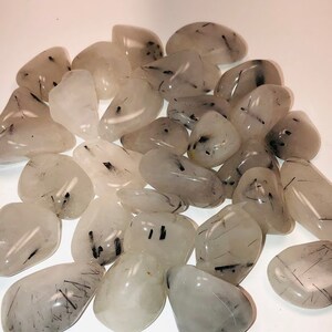 Puede incluir: Una colecci&oacute;n de piedras de cuarzo transparente pulidas con inclusiones de turmalina negra. Las piedras son lisas y pulidas, con una variedad de formas y tama&ntilde;os.