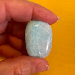 May include: A smooth, light blue and white aquamarine tumbled stone. The stone is oval shaped and has a polished finish.