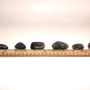 May include: Six black and brown tumbled stones of varying sizes and shapes, lined up on a wooden ruler with inch markings.