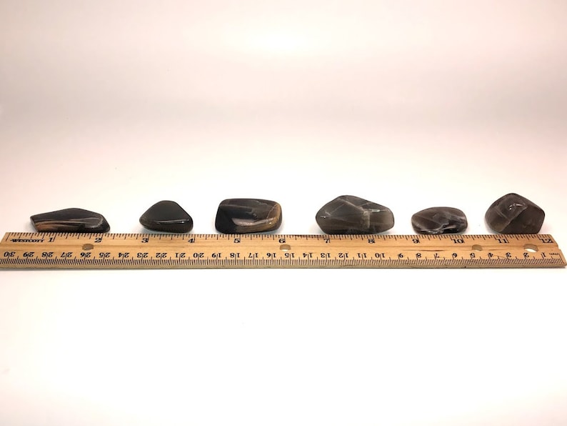 May include: Six black and brown tumbled stones of varying sizes and shapes, lined up on a wooden ruler with inch markings.