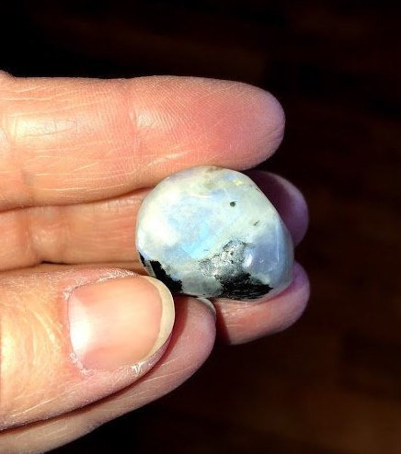 May include: A smooth, white, and iridescent moonstone with black inclusions. The stone is held in a hand.