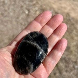 May include: A smooth, black obsidian palm stone with silver and white streaks. The stone is oval shaped and has a glossy finish.