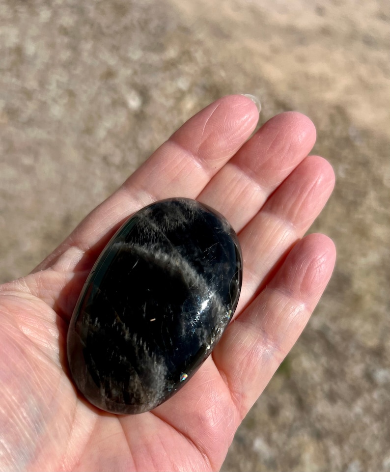 May include: A smooth, black obsidian palm stone with silver and white streaks. The stone is oval shaped and has a glossy finish.