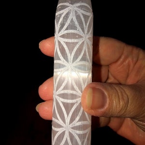May include: A white selenite wand with a carved flower of life design. The wand is about 6 inches long and is held in a hand.