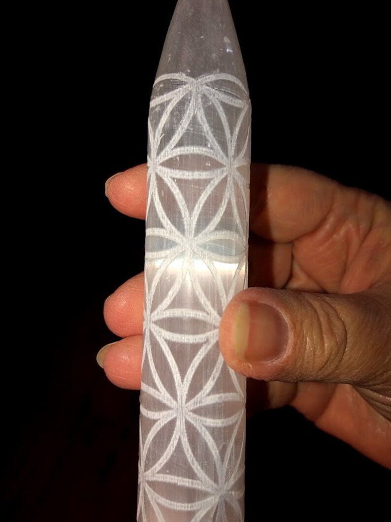 May include: A white selenite wand with a carved flower of life design. The wand is about 6 inches long and is held in a hand.