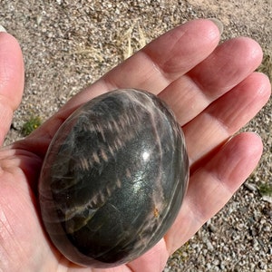 May include: A smooth, polished, dark gray and pink sunstone sphere with iridescent shimmer. The sphere is about the size of a golf ball.