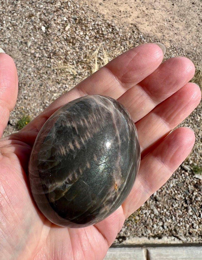 May include: A smooth, polished, dark gray and pink sunstone sphere with iridescent shimmer. The sphere is about the size of a golf ball.