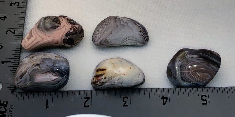 May include: Five agate stones with different patterns and colors, including pink, gray, and brown. The stones are arranged on a white surface with a ruler for scale.