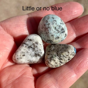 May include: Three tumbled stones with a white and gray speckled pattern. The stones have a light blue hue, with one stone having a more pronounced blue color. The text "Little or no blue" is visible above the stones.