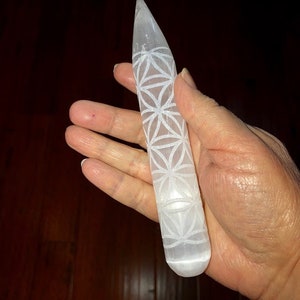 May include: White selenite crystal wand with a carved flower of life design. The wand is about 6 inches long and has a rounded tip.