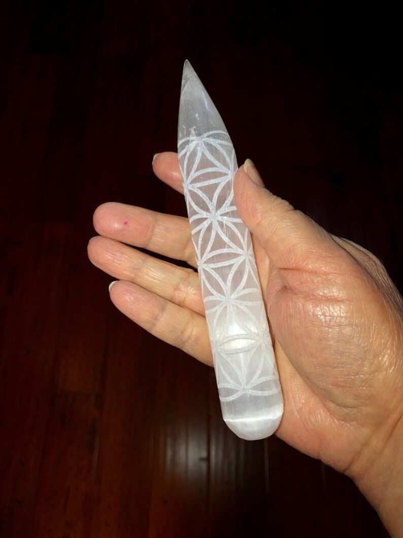 May include: White selenite crystal wand with a carved flower of life design. The wand is about 6 inches long and has a rounded tip.