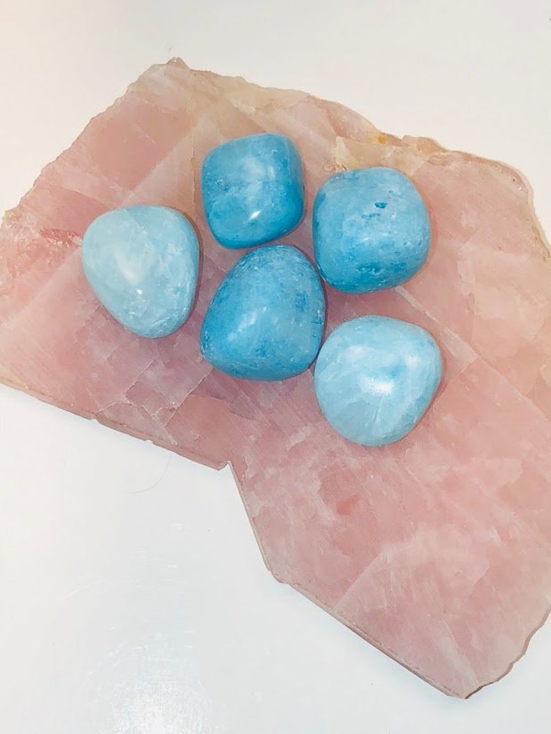 May include: Five smooth, light blue tumbled stones on a pink, textured surface. The stones are various sizes and shapes.