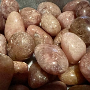May include: A collection of tumbled rose quartz stones in various shades of pink and brown. The stones are smooth and polished, with a translucent appearance.
