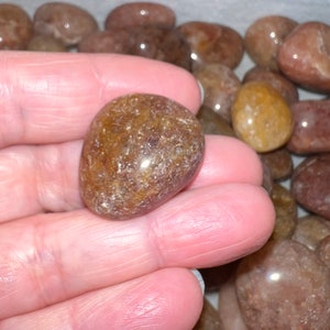 May include: A close-up of a hand holding a smooth, brown, translucent tumbled stone. The stone has a slightly cloudy appearance with flecks of white and brown throughout.